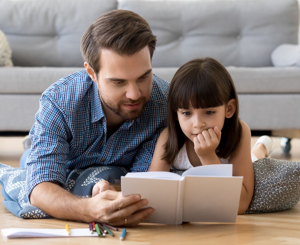 Cute little girl listening to dad reading fairy tale lying on warm floor together, caring father holding book teaching child daughter spending time with focused kid, family hobbies activities at home (Cute little girl listening to dad reading fairy ta